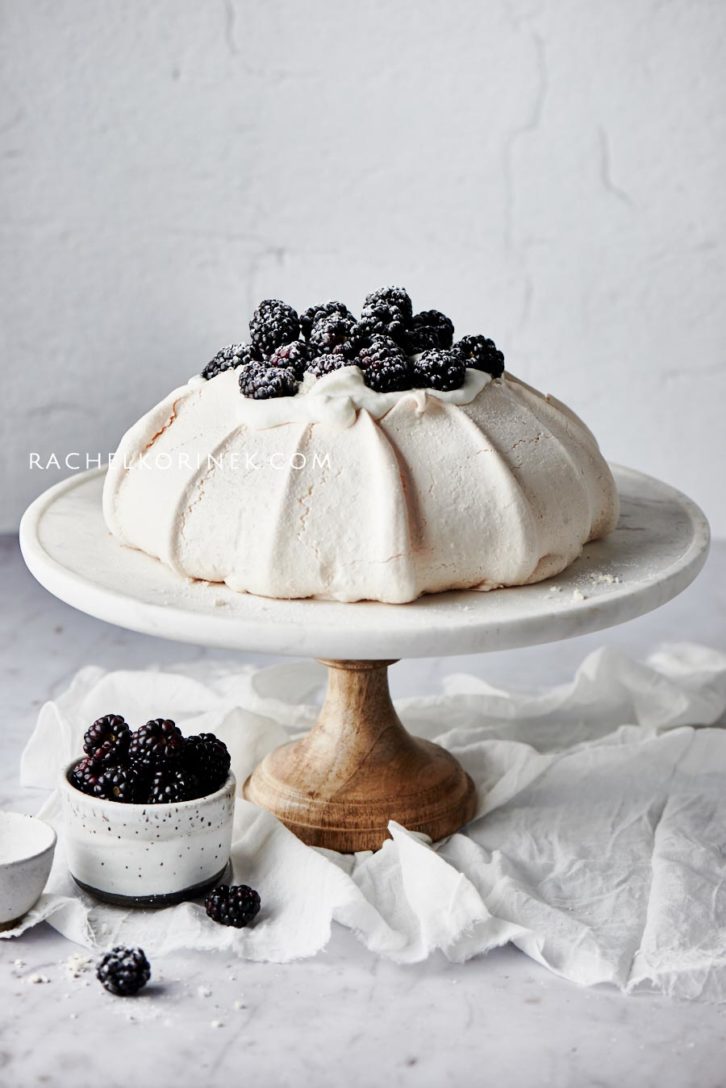 photos of a pavlova with blackberries on top. On a cake stand with a napkin underneath. There isn't any colour to this photo so it appears black and white even thought it's real food!