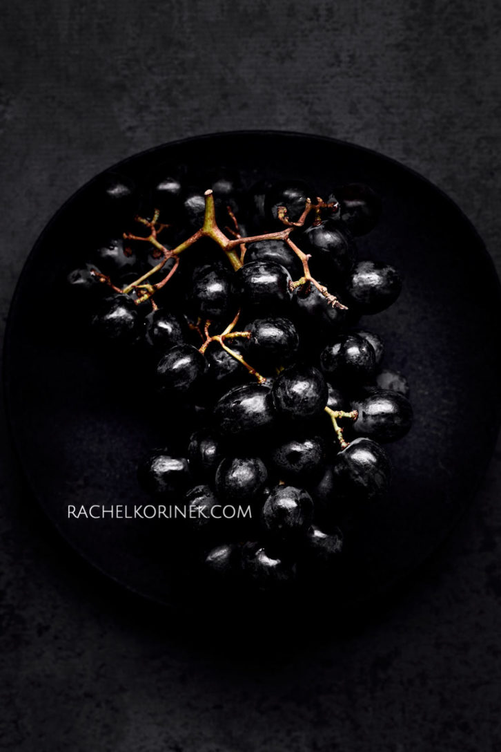 Dark black grapes sitting on a black plate. There isn't any colour to this photo so it appears black and white even thought it's real food!