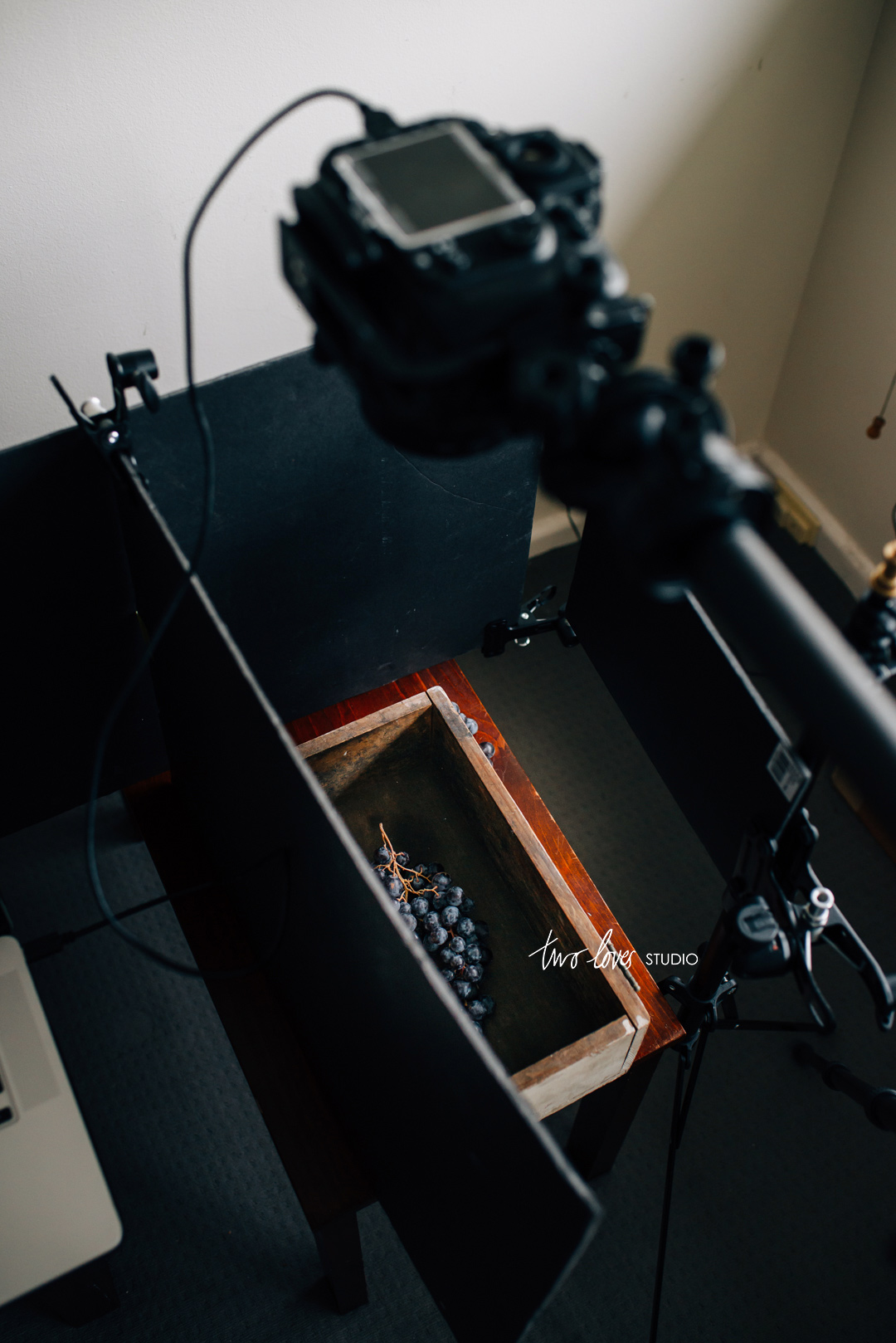 Behind the scenes shot of purple grapes in a wooden box on a dark background with a camera above. 