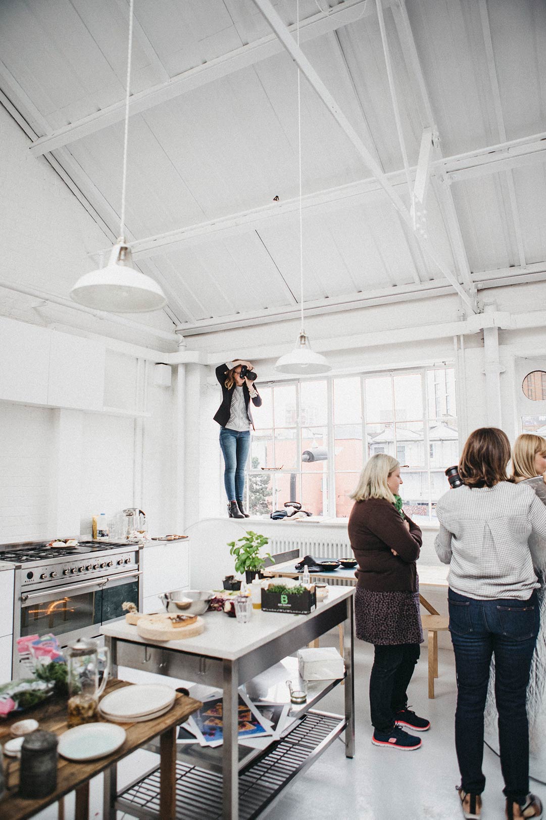 Food photography workshop with people standing around watching a demonstration. A photographer is standing on the window sill to capture to behind the scenes.