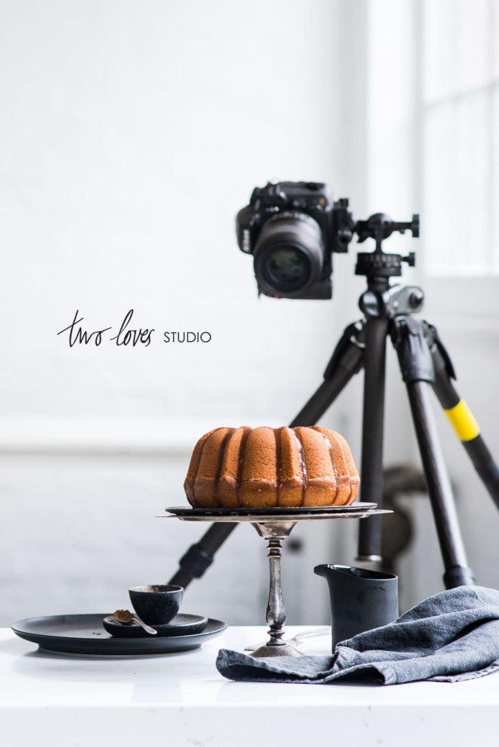 Camera on tripod shooting a bundt cake on a cake stand in a white studio. Behind the scenes food photography setup.