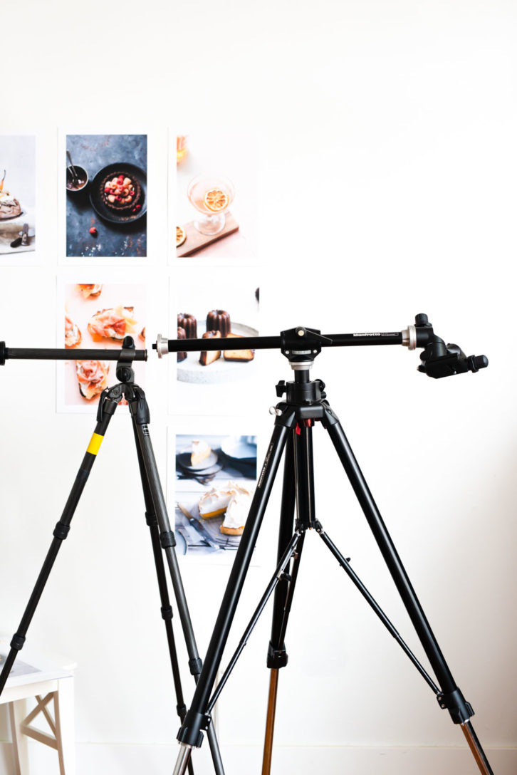 A tripod in full extension with a white wall behind it that has images of food pinned to the wall. 