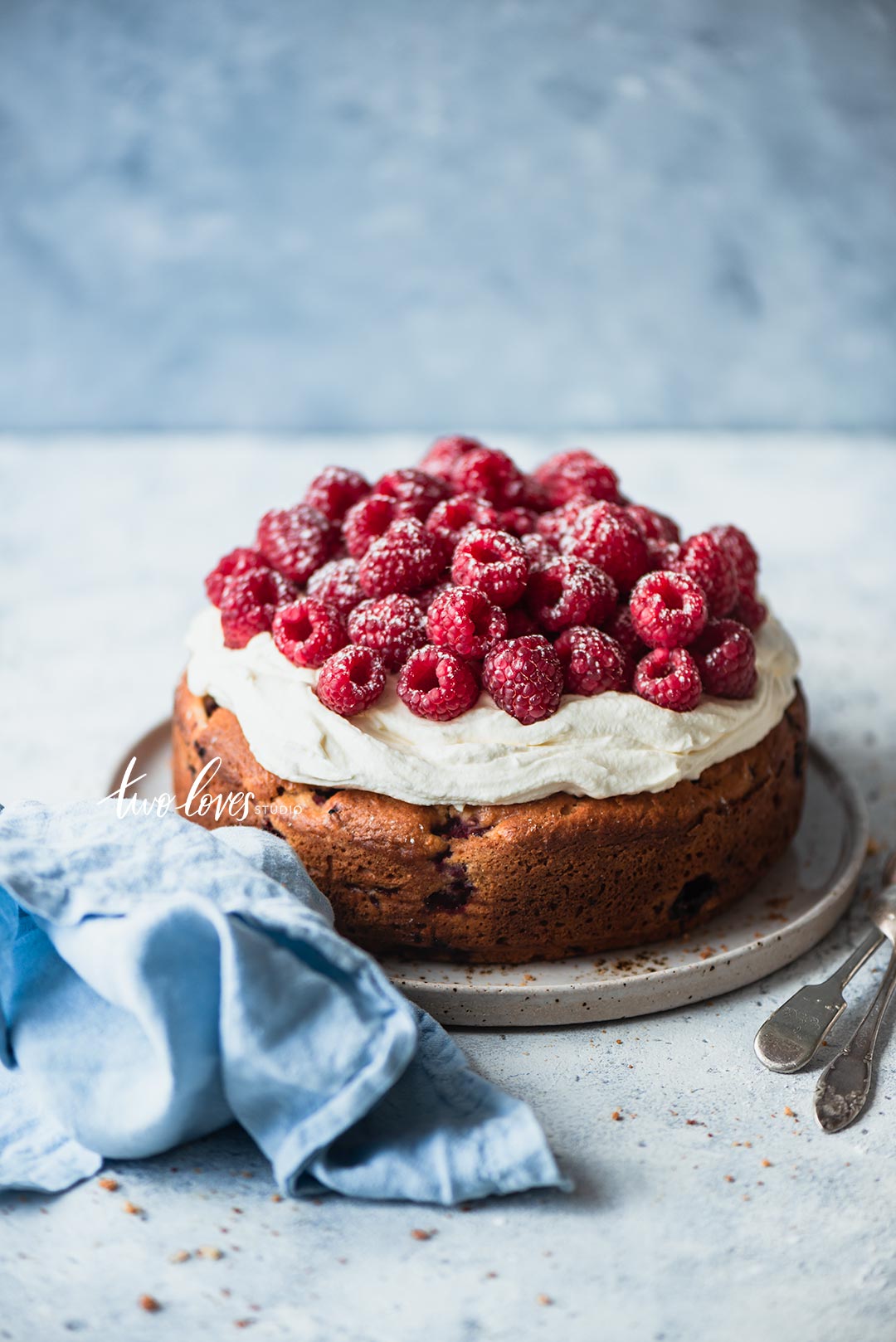 A close up photo of a cake with creme cheese topping and raspberries dusted with icing sugar and a blue napkins. 