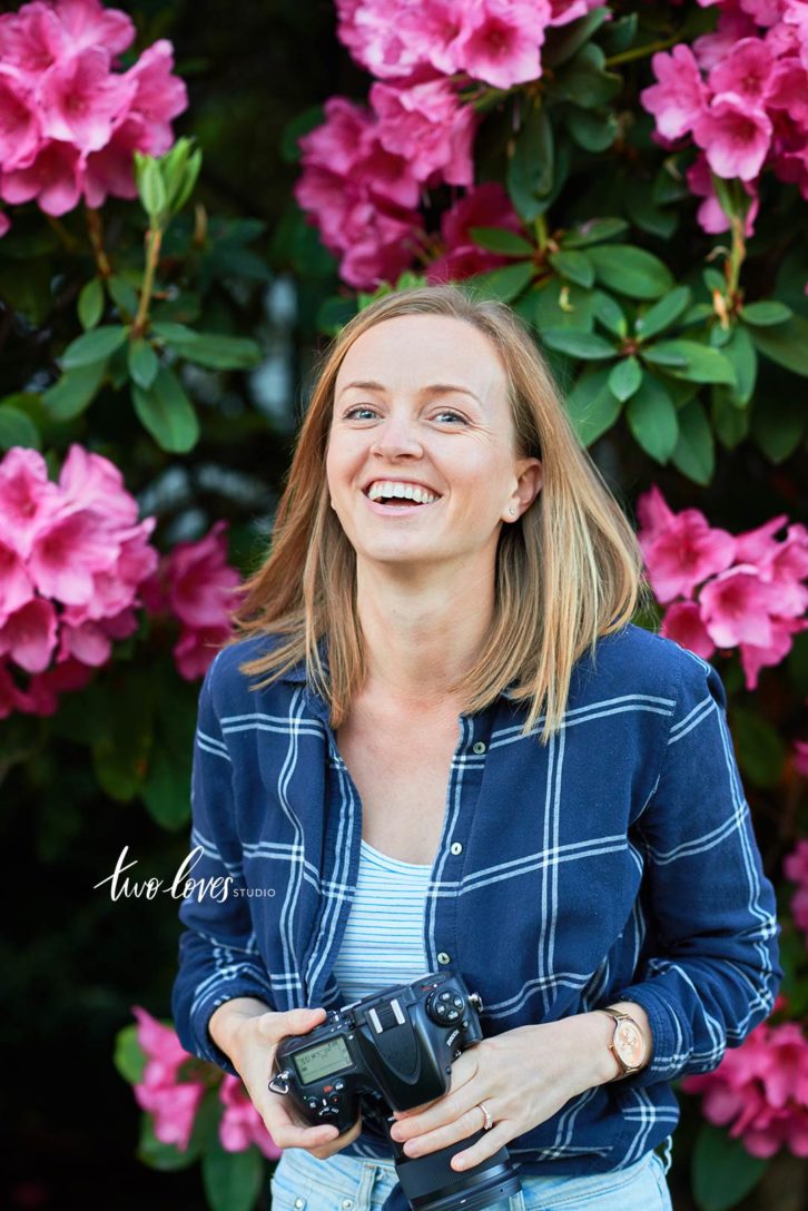 Rachel smiling in front of a bush with pink flowers, holding a camera. 