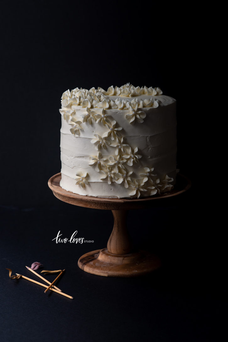 A cake stand with a four teared cake with a white frosting and flowers piped along the side 