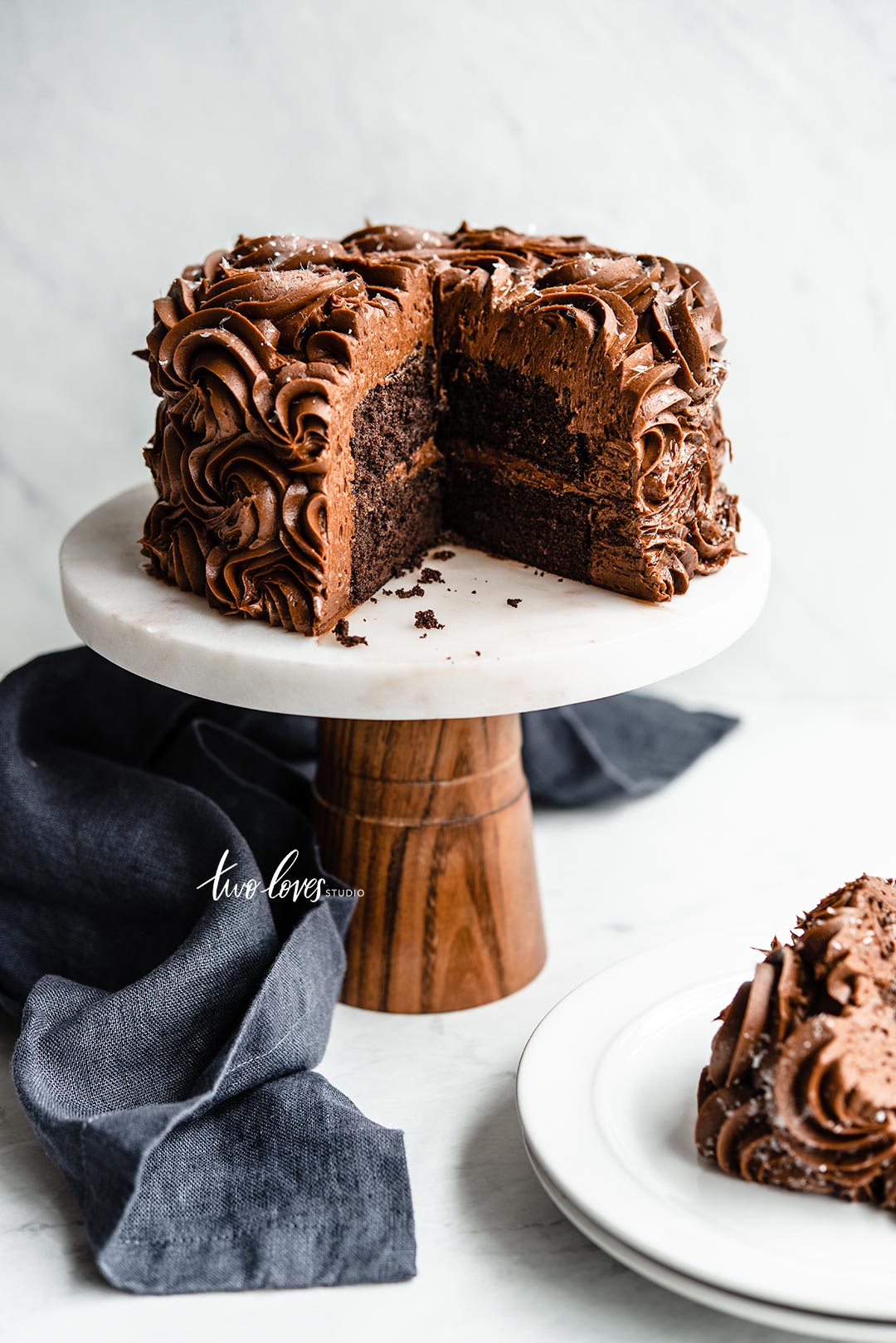 Chocolate cake on a white cake stand, slice of cake and a blue napkin on the side. Demonstrating a narrow lens for straight lines.