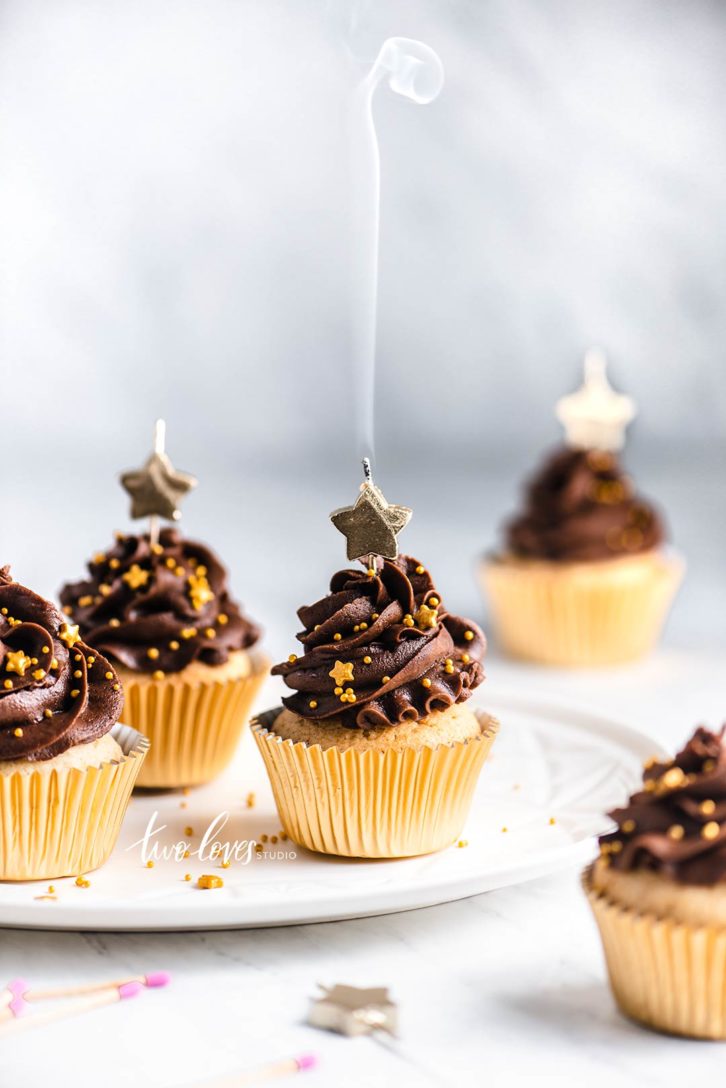 A bunch of cupcakes with gold cupcake holders and high chocolate topping. Star candles on top. 