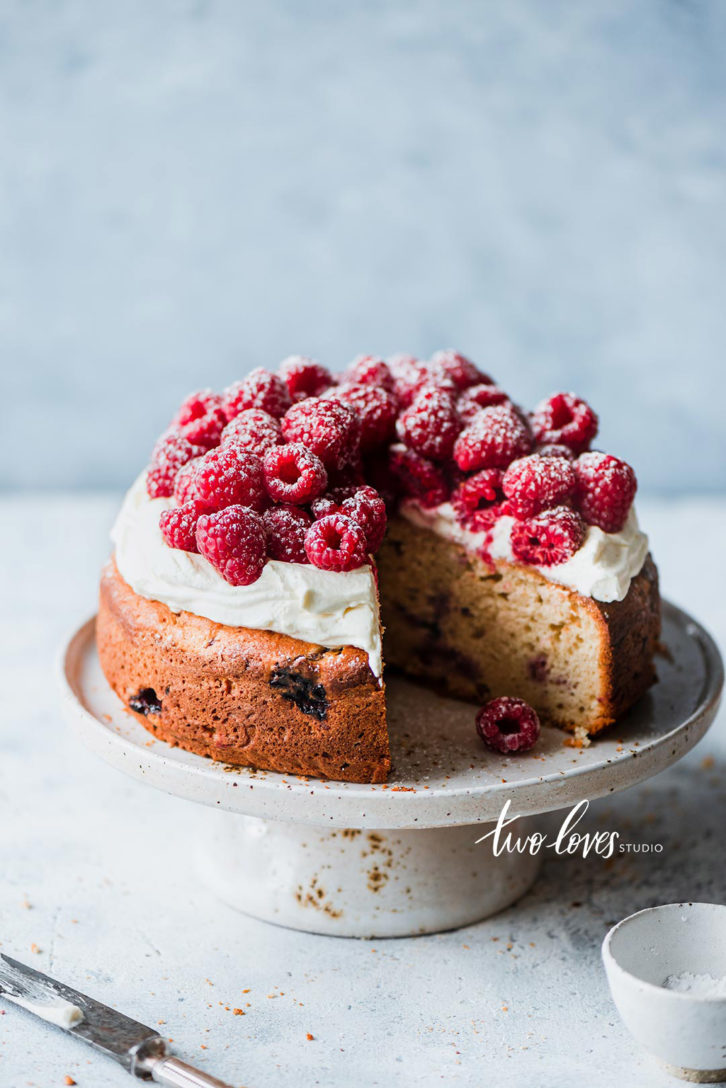 A cake stand with a cake topped with cream cheese and raspberries. 