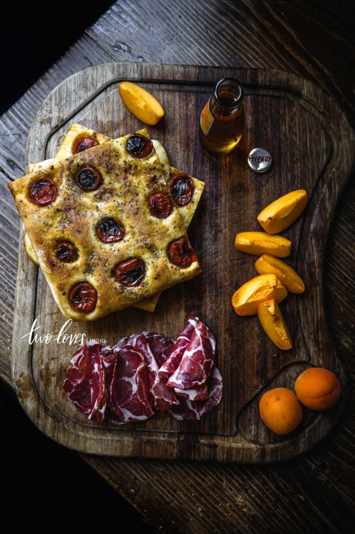 Wooden chopping board with tomato pizza, cured meat and slices of fruit.