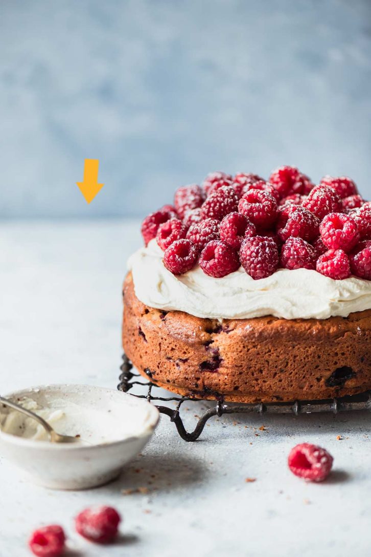 Circular cake sitting on a wire cooling rack, with a cream cheese frosting on top and raspberries to finish.