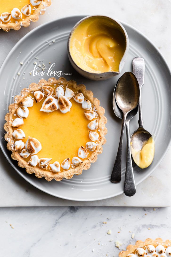 Lemon meringue tart wreaths on a grey plate with silver spoons and a side pot of lemon tart filling.