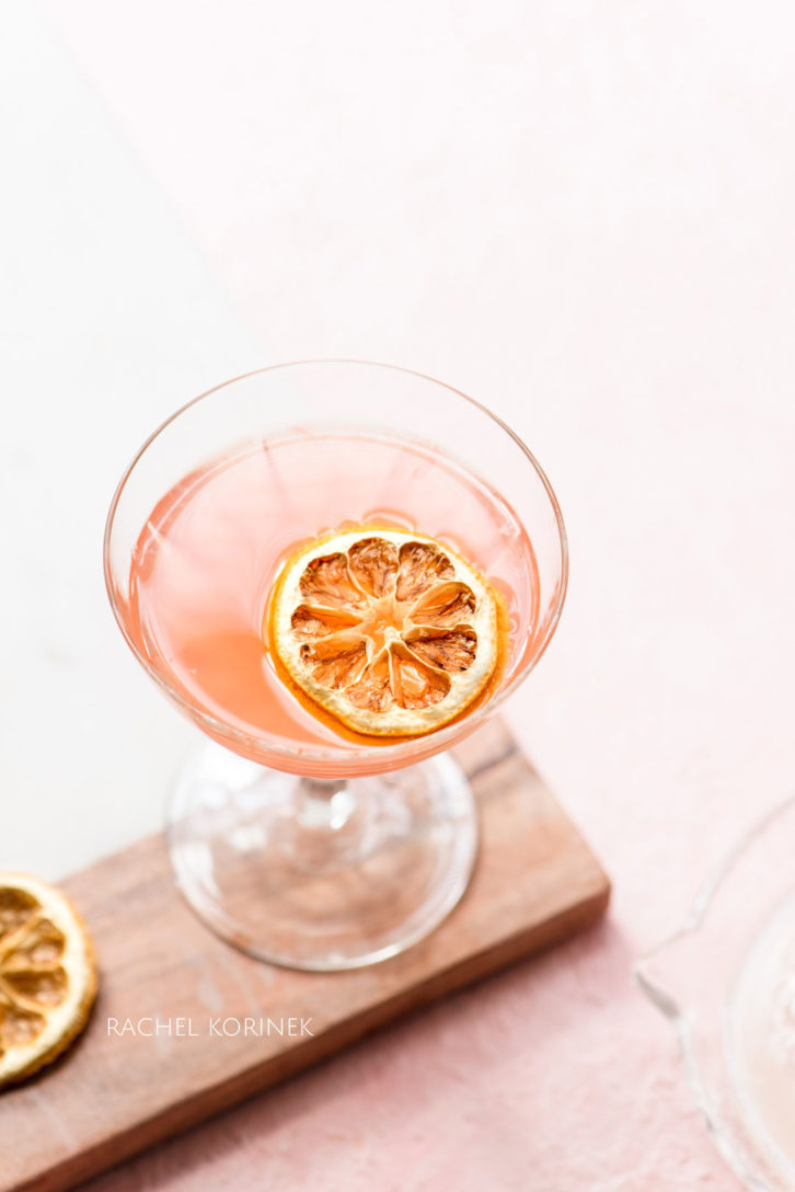 Pink cocktail in a classic cocktail glass, with a decorative dried lemon slice.