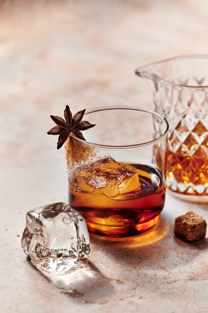 A whiskey glass filled with rich whiskey, a clear ice cube and garnished with raw sugar and a star anise.