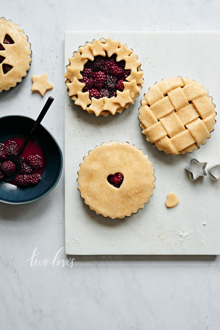 Four mini blackberry pies on a white background. Each pie has a unique lattice work design.