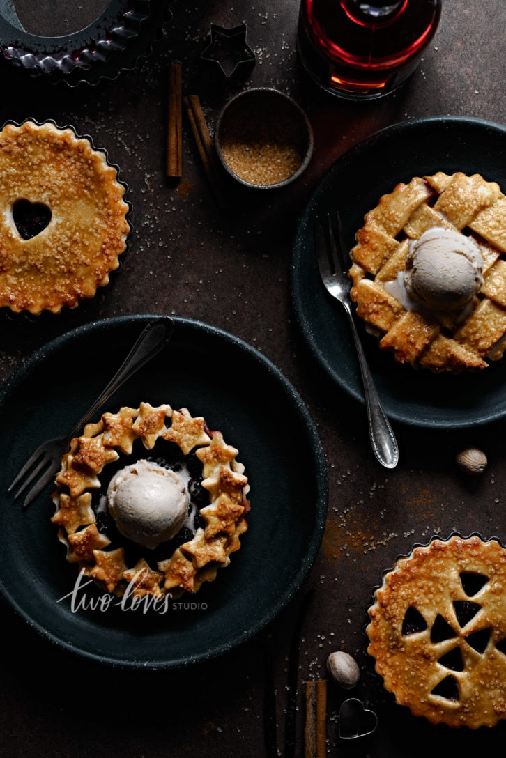 Four mini blackberry pies on a brown background. Each pie has a unique lattice work design. There is ice cream on top of two of the pies.