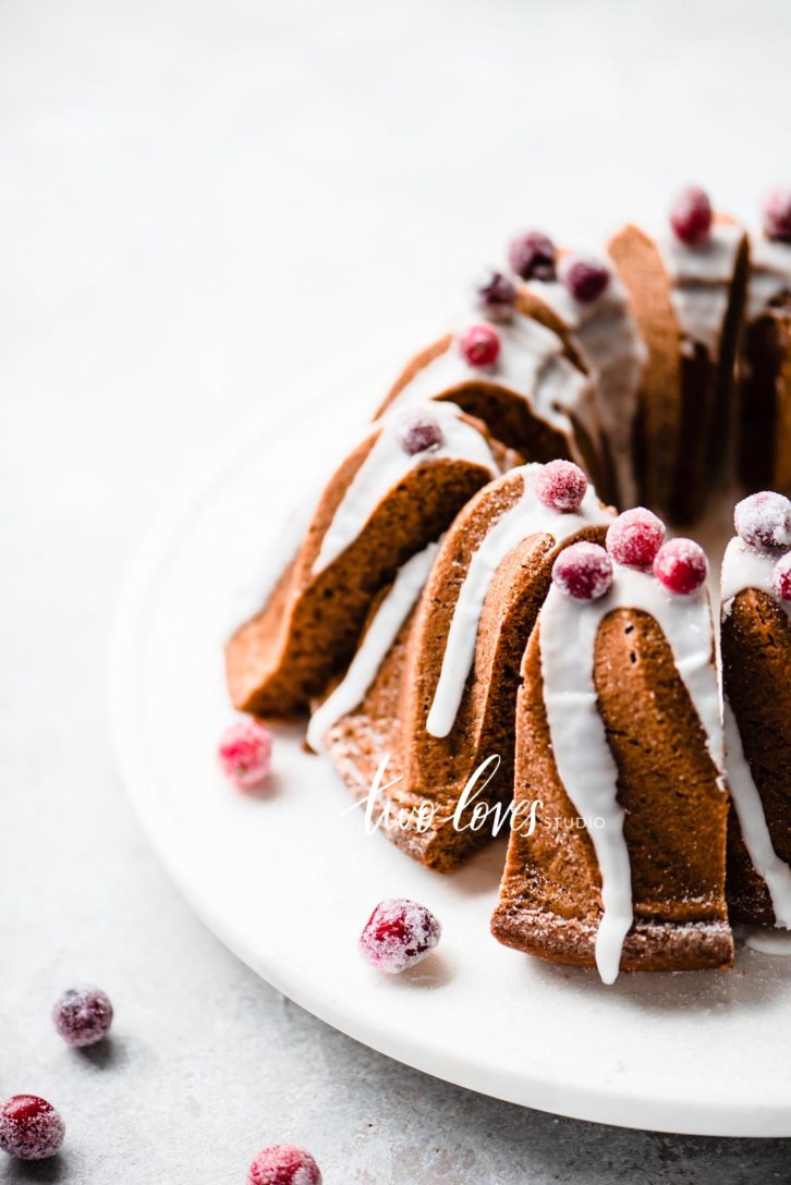 A gingerbread bundt cake cut into slices with icing drizzle and sugared cranberries.