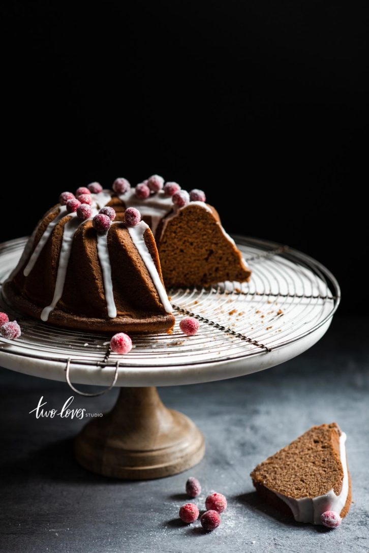 A gingerbread bundt cake cut into slices with icing drizzle and sugared cranberries on a cake stand.