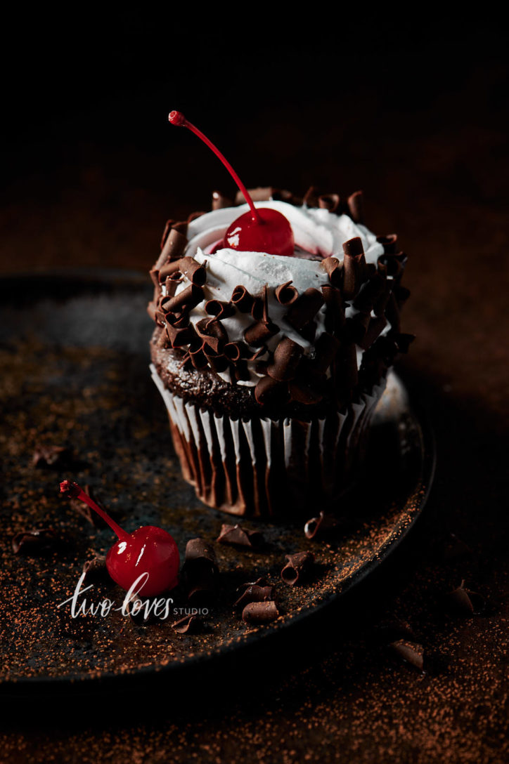 Black forest cupcake with a cherry on top. Sitting on a brown plate, with whipped cream and chocolate curls.