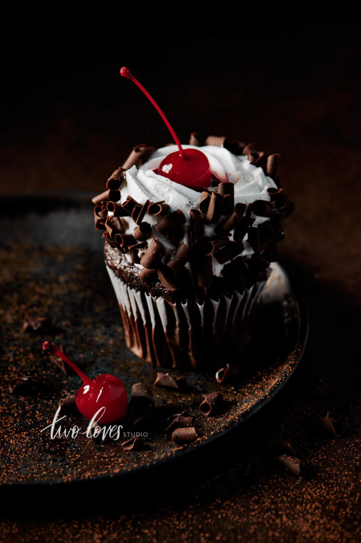 Black forest cupcake with a cherry on top. Sitting on a brown plate, with whipped cream and chocolate curls.