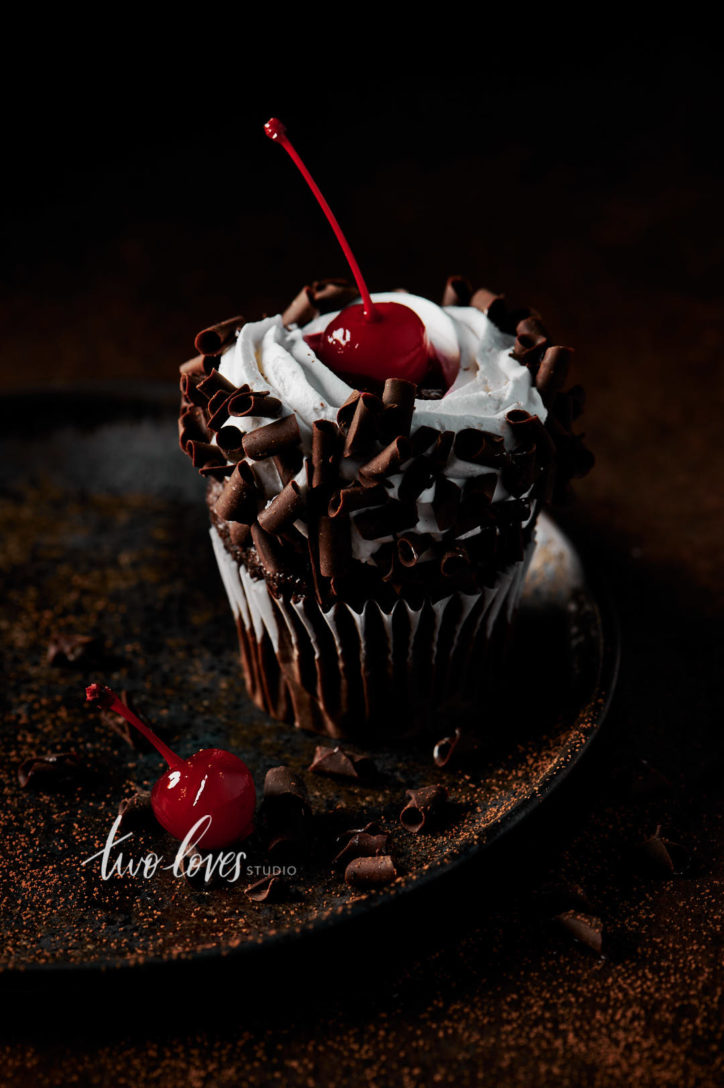 Black forest cupcake with a cherry on top. Sitting on a brown plate, with whipped cream and chocolate curls.