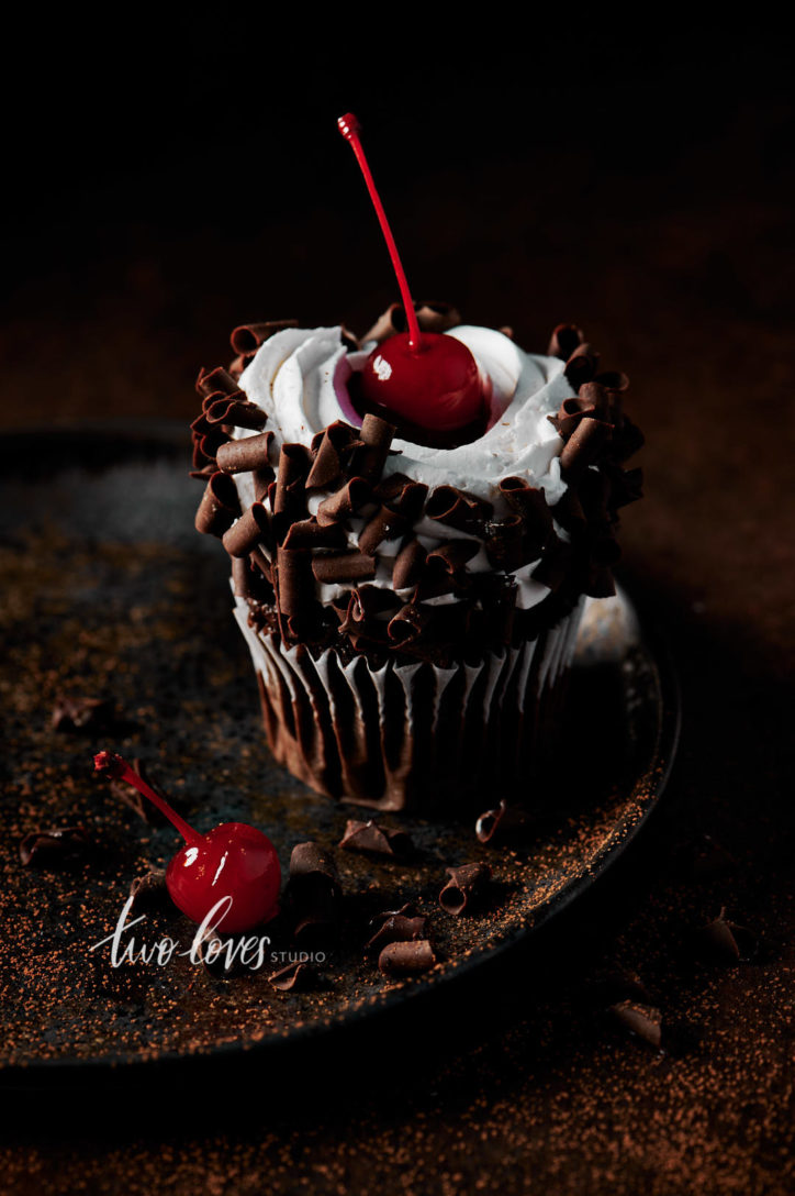 Black forest cupcake with a cherry on top. Sitting on a brown plate, with whipped cream and chocolate curls.