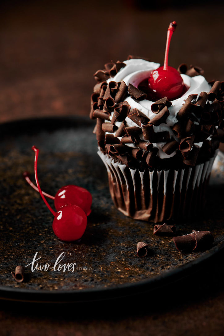 Black forest cupcake with a cherry on top. Sitting on a brown plate, with whipped cream and chocolate curls.