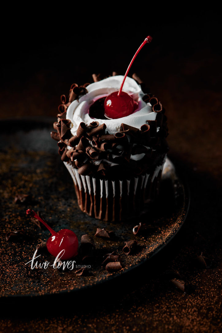 Single chocolate cupcake with a cherry on top on a dark coloured dish on a chocolate coloured backdrop. 