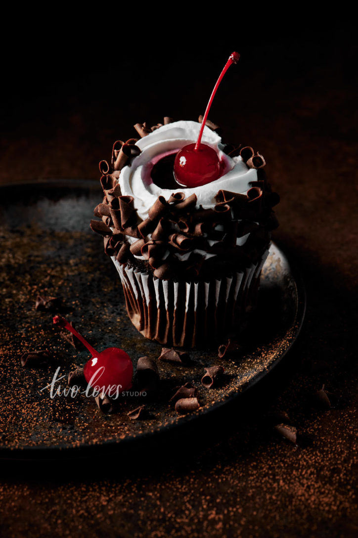 Black forest cupcake with a cherry on top. Sitting on a brown plate, with whipped cream and chocolate curls.