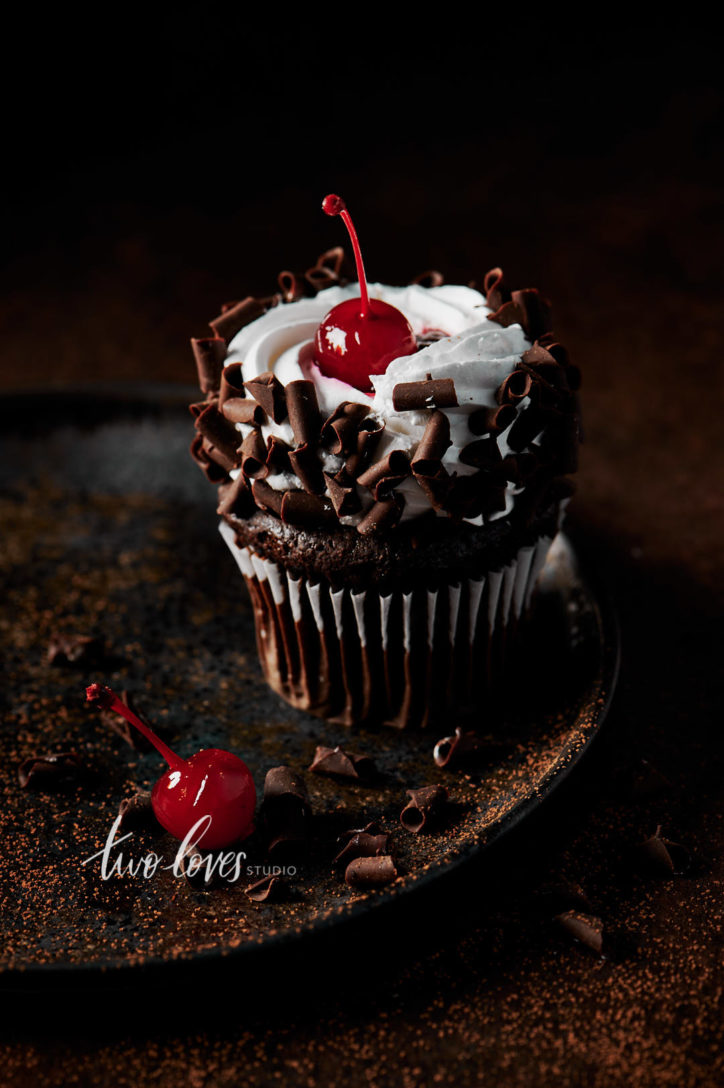 Black forest cupcake with a cherry on top. Sitting on a brown plate, with whipped cream and chocolate curls.