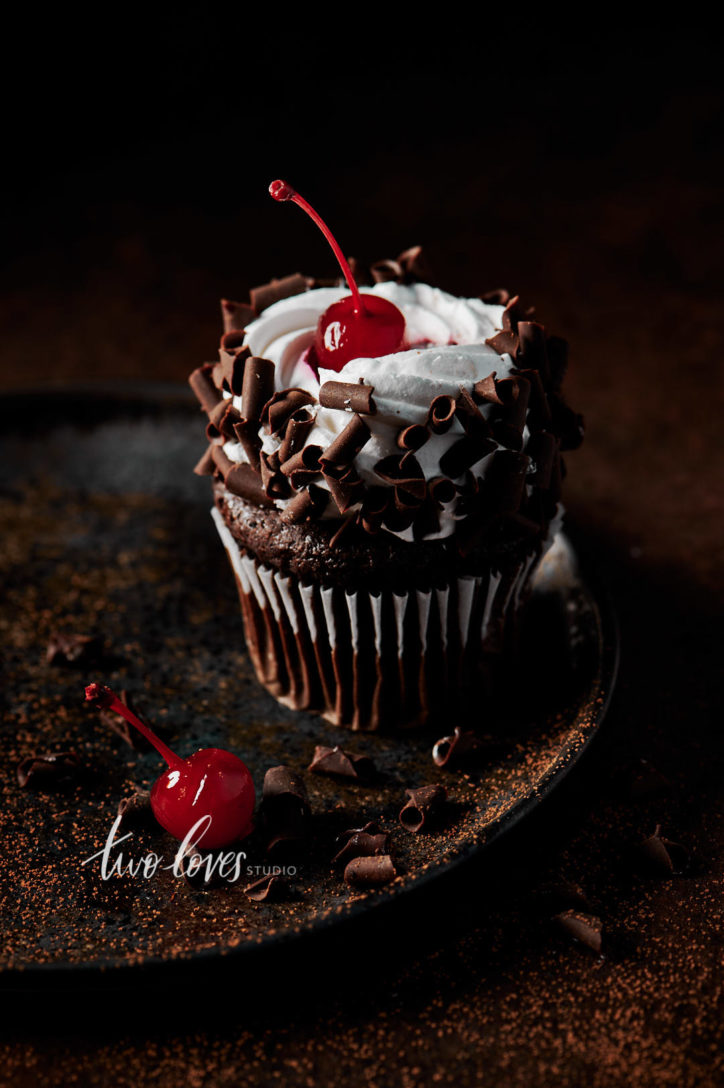 Black forest cupcake with a cherry on top. Sitting on a brown plate, with whipped cream and chocolate curls.