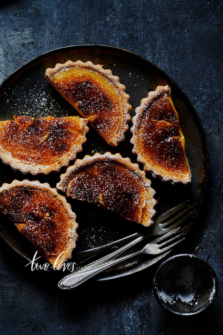 lemon tarts that have been bruleed on  a dark plate with three forks.