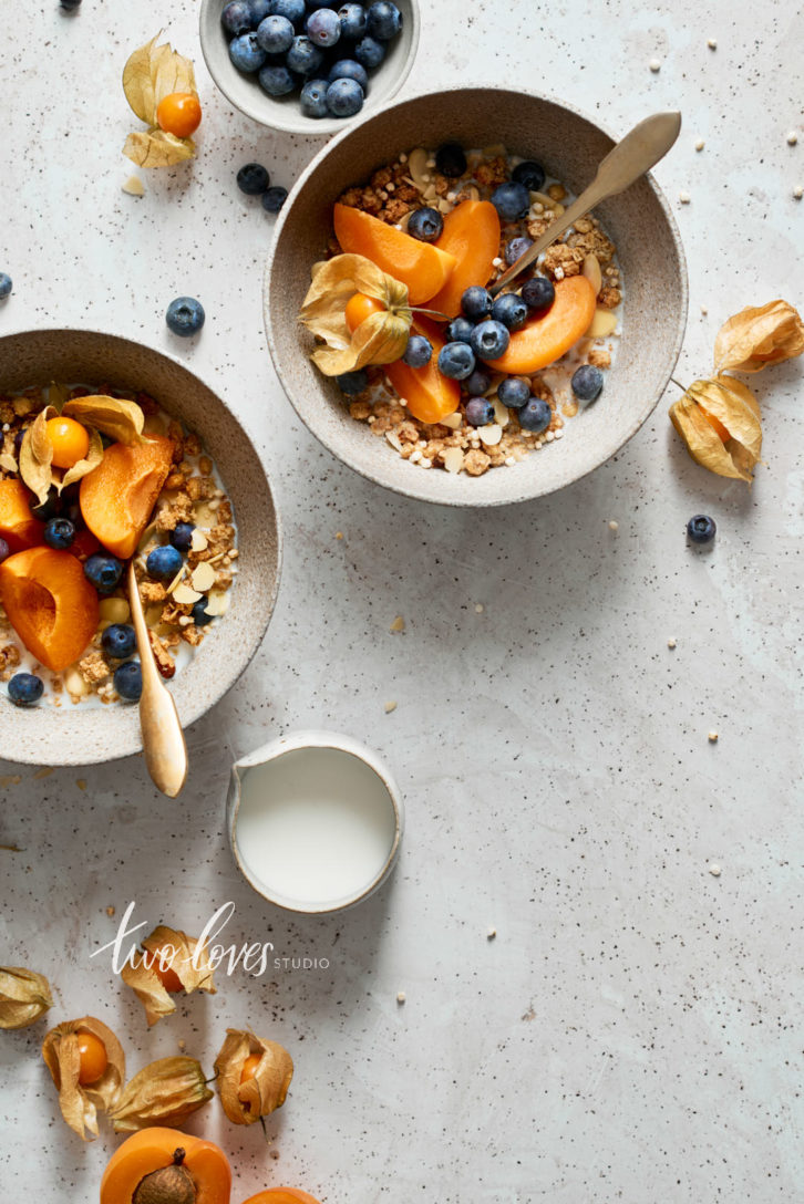 Two bowls of granola with blueberries and sliced apricots with golden spoons.