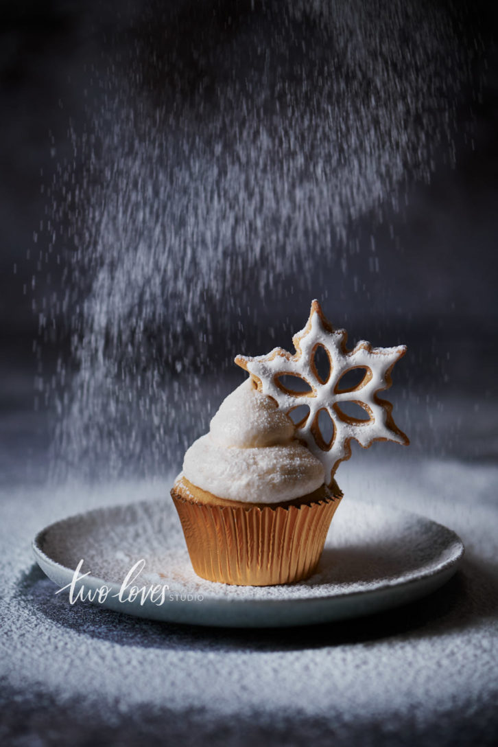 A white cupcake in a gold wrapper with a star shaped cookie and falling icing sugar. Showing the best camera settings for food photography when you want to blur motion.