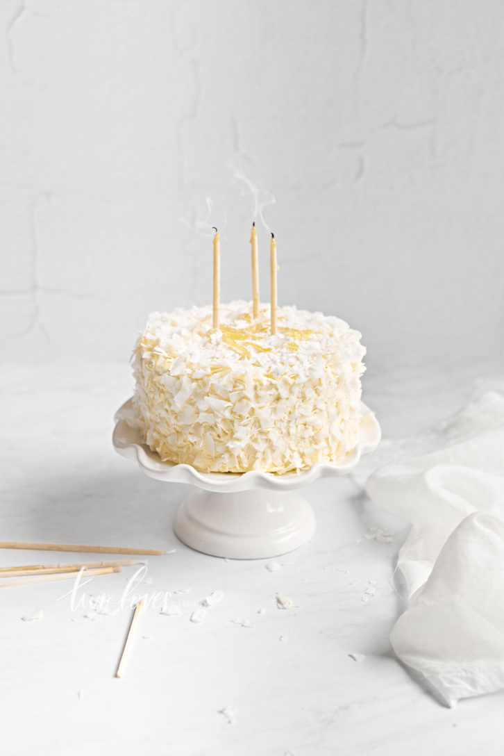 A white coconut cake on a white cake stand on a white background. Three beeswax candles on the top with smokey details.