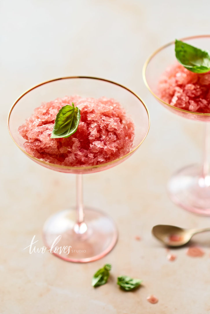 Soft pink, white background with 2 pink martini glasses filled with watermelon slushy and a basil leaf.