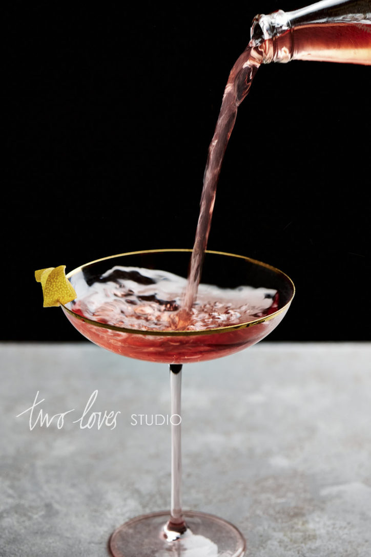 A large pink cocktail glass with a lemon garnish with a pink soda being poured in from above. The camera settings reveal how to blur the motion in this image.