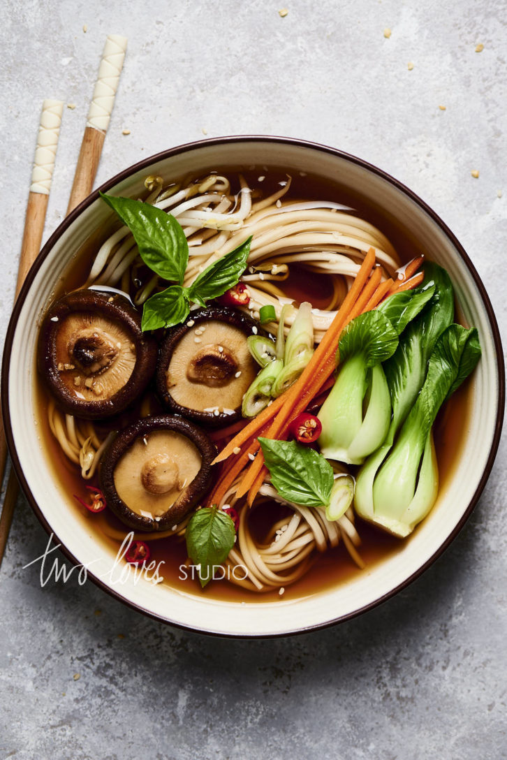 A bowl of pho with noodles, baby bok choy, julienne carrots, shiiatkes, thai basil and chillies. Scattered with sesame seeds and a pair of chopsticks. Showing the camera settings for food photography.