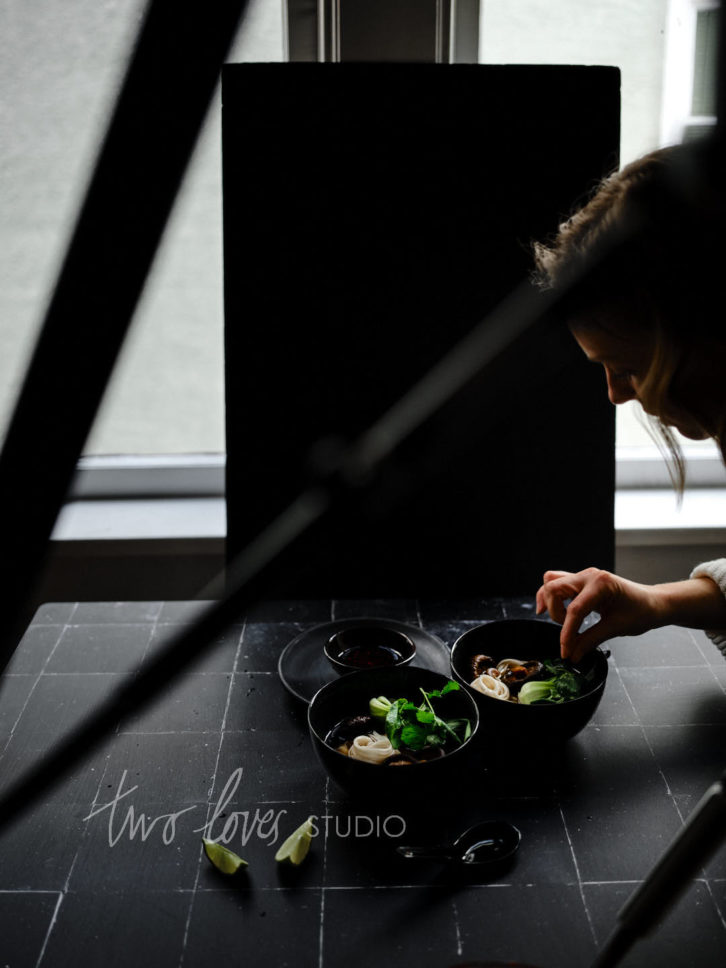 Two bowls of ramen in black bowls on a black background with a hand making adjustment. 