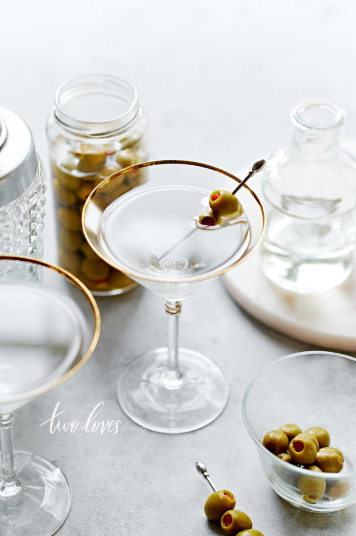 A dirty martini with a two olives on a cocktail pick. With some cocktail making accessories in the foreground and background.