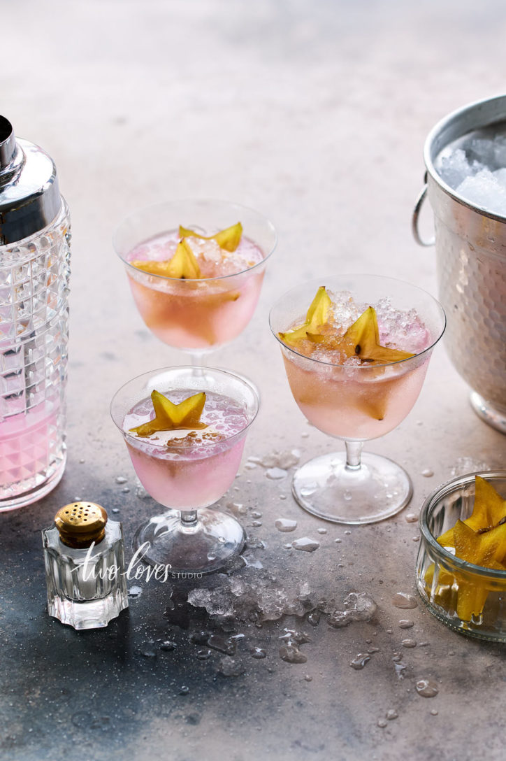 An icy shot a gin and tonic in 3 small glasses with star fruit. A bucket of ice and a vintage cocktail shaker in the background.