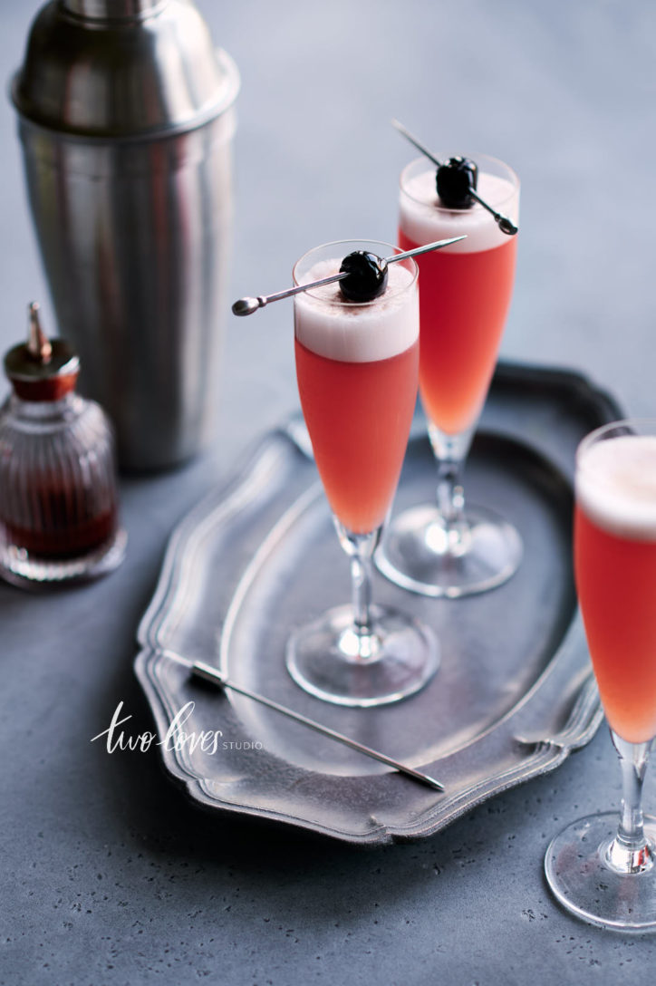 Gin blood orange sour in two mini champagne glasses with a cherry on top on a cocktail pick.