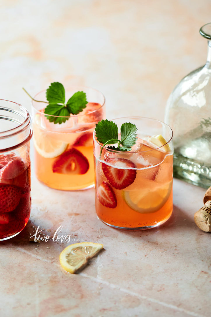 Two short tumbler glasses with sangria, strawberries and lemon slices on a pale back drop. 