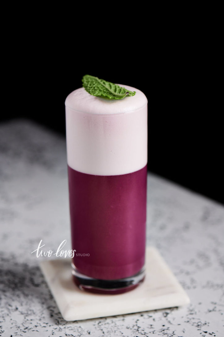 Magenta cocktail in a tall glass with a thick egg white dry shake cocktail foam and a single mint leaf on top.  