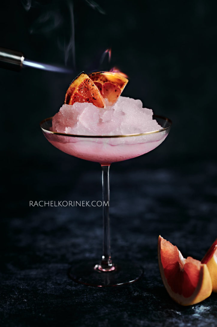 Pink frozen cocktail on a dark background with a chard orange slice as the cocktail garnish.
