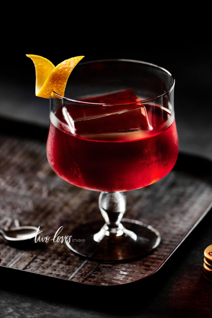 Drink photography of a single red cocktail drink in a glass with a single piece of ice and a lemon garnish slice. 