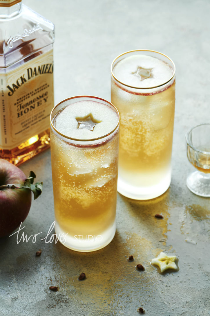 Jack Daniel cocktails on a grey backdrop in two tall glasses with apple circle garnishes.