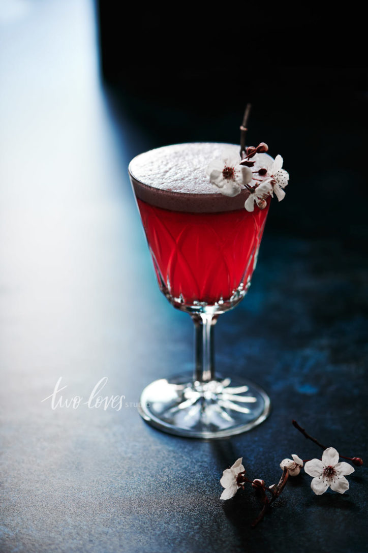 Single cocktail with a cherry blossom cocktail garnish on a dark blue background. 