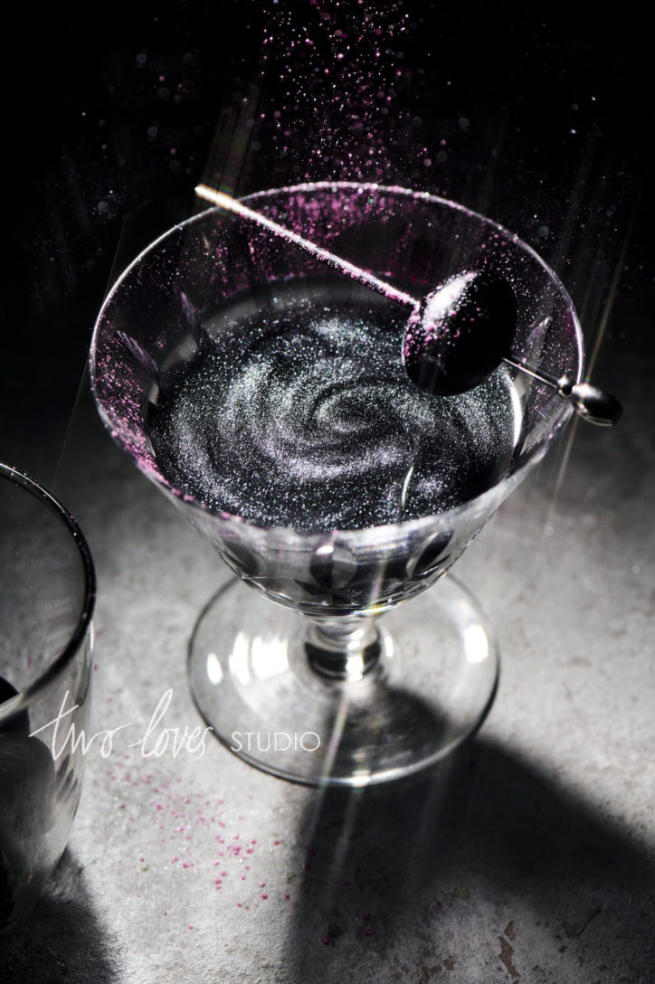 Drink photo of a midnight black cocktail with edible glitter on top and a black olive garnish. 