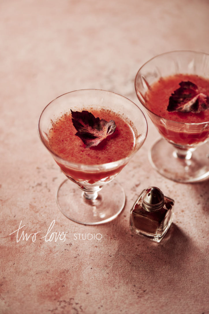 Two red cocktails with a single maple leaf garnish and a mini spice shaker on the side.