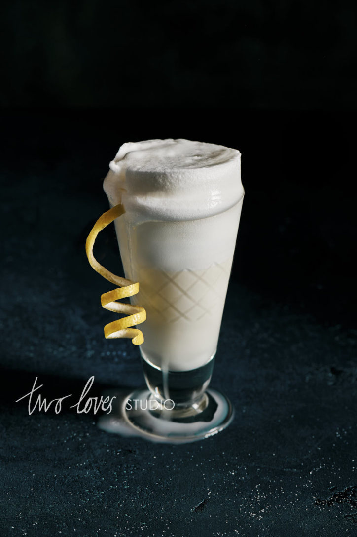 Black background with a single tall glass filled with a ramos fizz and a lemon twist.